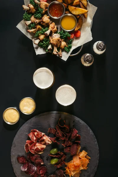 Вид зверху на келихи пива, асорті м'яса та смаженої картоплі з куркою на чорному — стокове фото