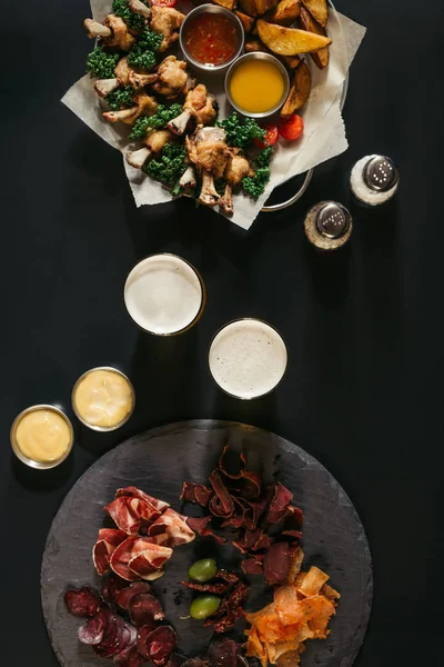 Vue du dessus des verres de bière, de la viande assortie et des pommes de terre frites avec du poulet sur noir — Photo de stock