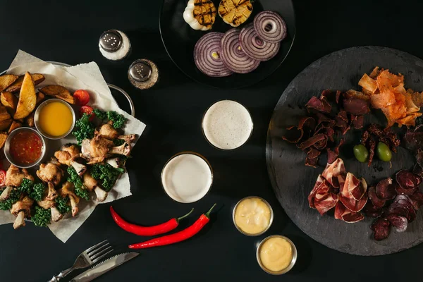 Vista superior de vasos de cerveza, verduras, papas al horno con pollo asado, varias salsas y carne artesanal en negro - foto de stock