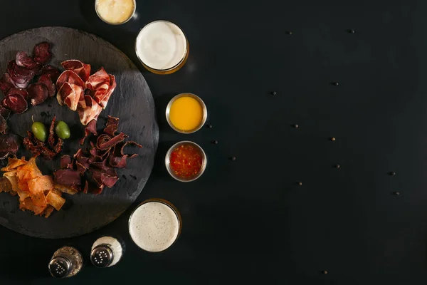 Vue de dessus de délicieuses tranches de viande assortie, verres de bière, sauces et épices sur noir — Photo de stock