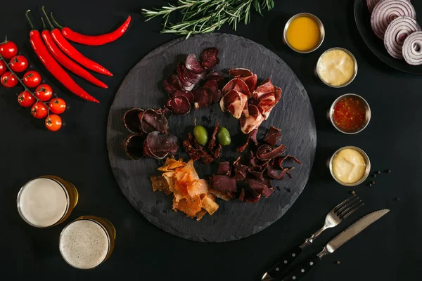 Gourmet assorted meat on slate board, various sauces, vegetables and glasses of beer on black — Stock Photo