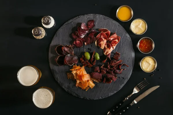 Вид зверху на гастрономічне м'ясо на дошці для шиферу, різні соуси та келихи пива на чорному — стокове фото