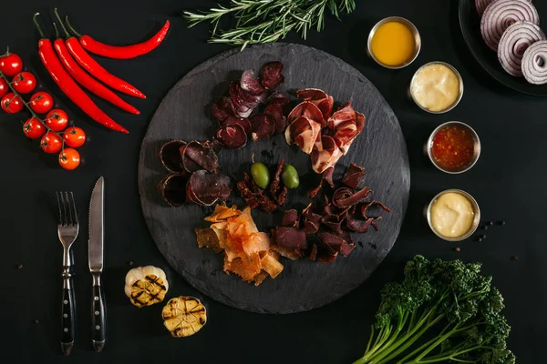 Вид на изысканные сорта мяса на доске, различные соусы и овощи на черном — стоковое фото