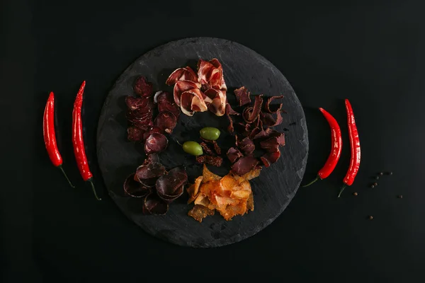 Top view of gourmet assorted meat with olives on slate board and chili peppers on black — Stock Photo