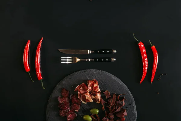 Top view of gourmet assorted meat on slate board, fork with knife and chili peppers on black — Stock Photo