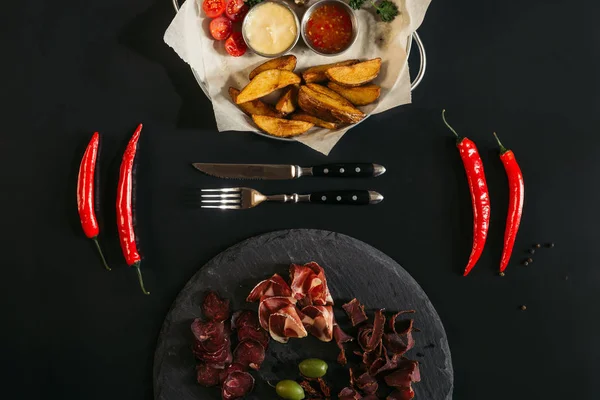 Top view of baked potatoes with sauces, chili peppers, fork, knife and gourmet assorted meat on slate board on black — Stock Photo