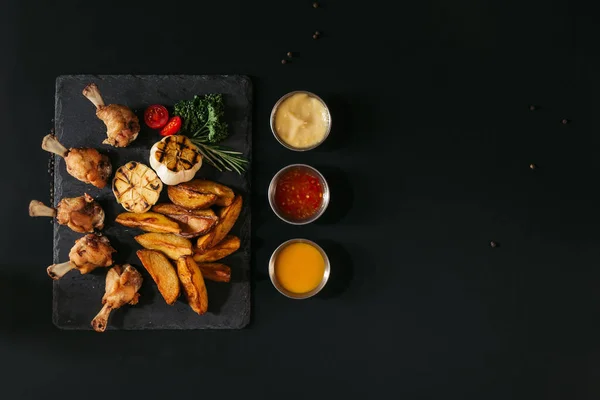 Vista dall'alto di gustose patate al forno con aglio alla griglia e pollo, erbe e salse su nero — Foto stock