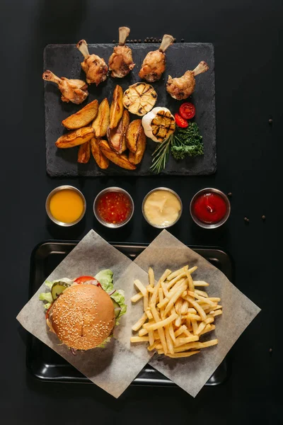 Vista superior de papas fritas con deliciosa hamburguesa en bandeja, salsas surtidas y pizarra con patatas asadas, verduras a la parrilla y alas de pollo en negro - foto de stock