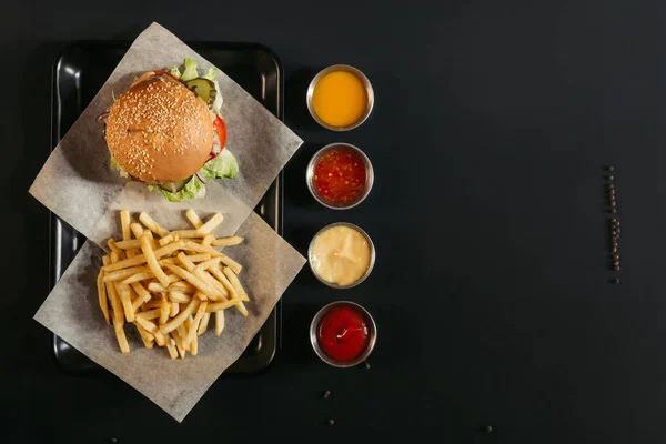 Vista superior de papas fritas con deliciosa hamburguesa en bandeja y salsas surtidas en negro - foto de stock