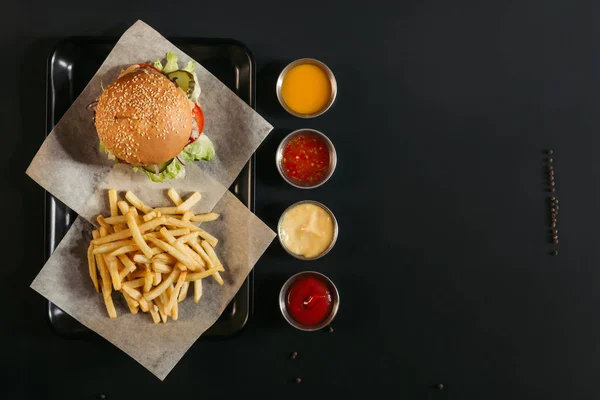 Top view of french fries with delicious burger on tray and assorted sauces on black — Stock Photo
