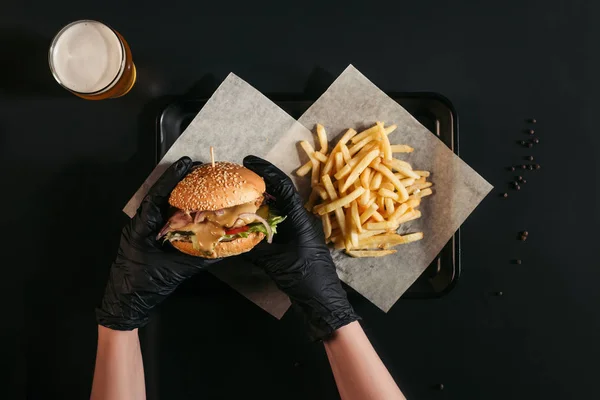 Cropped shot of person in gloves holding tasty burger above tray with french fries and glass of beer on black — Stock Photo