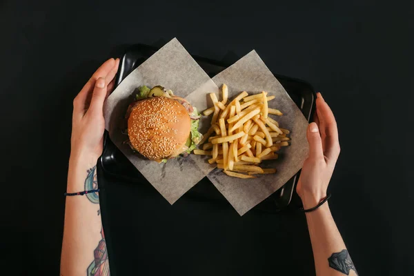 Cropped shot of human hands holding tray with delicious burger and french fries on black — Stock Photo