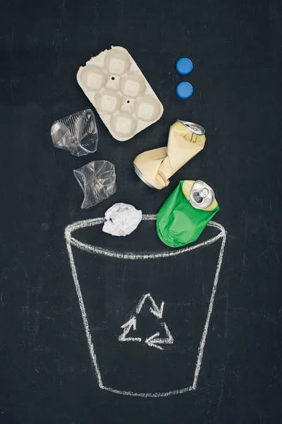 Different types of trash falling into drawn trash bin with recycle sign on chalkboard — Stock Photo