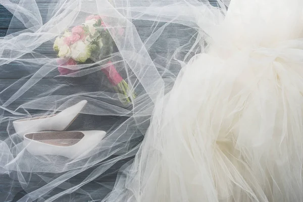 Vista superior de par de zapatos, vestido de novia y ramo de madera sobre mesa azul oscuro - foto de stock