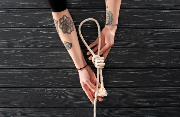 Partial view of female tattooed hands with nautical rope with knot on dark wooden surface — Stock Photo