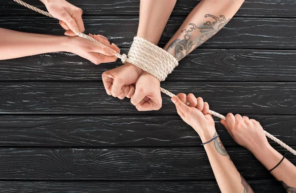 Partial view of women tying rope around males hands on dark wooden tabletop — Stock Photo