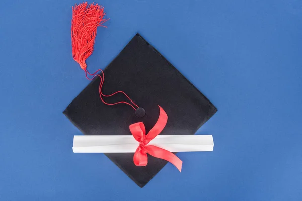 Graduation hat and diploma with red ribbon isolated on blue — Stock Photo