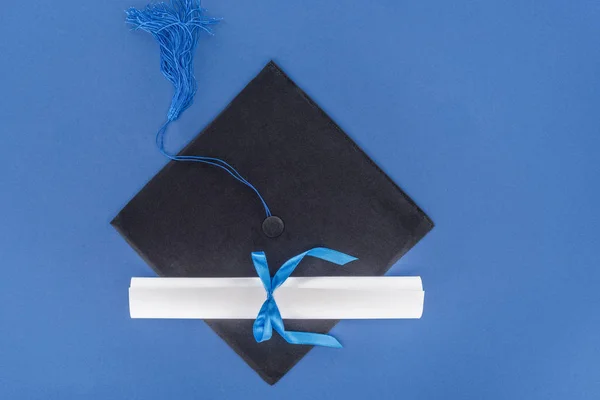 Graduation hat and diploma with blue ribbon isolated on blue — Stock Photo
