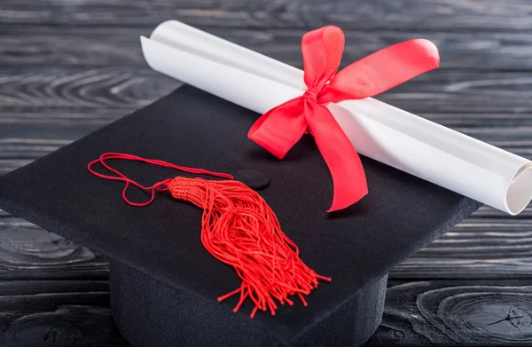 Graduation hat and diploma with red ribbon on wooden table — Stock Photo