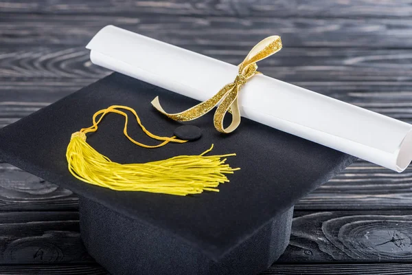 Graduation hat and diploma with yellow ribbon on wooden table — Stock Photo