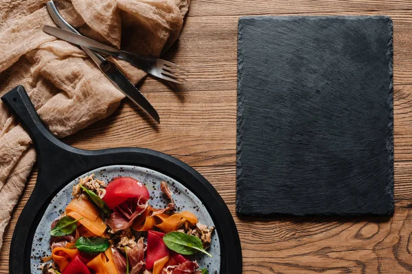 Салат с мидиями, корневыми овощами и хамоном, вилка с ножом и доска с чистого листа на деревянном столе — стоковое фото