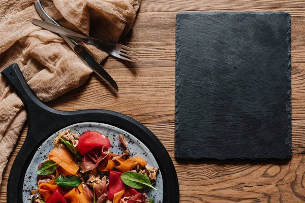 Salada com mexilhões, legumes de raiz e jamon, garfo com faca e tábua de ardósia em branco na mesa de madeira — Fotografia de Stock