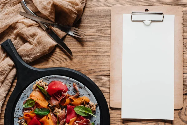 Salad with mussels, vegetables and jamon, fork with knife and blank paper on clipboard on wooden table — Stock Photo