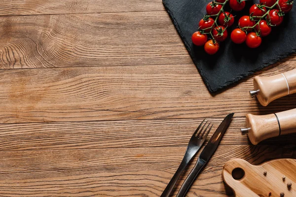 Vista superior de tomates maduros frescos en pizarra, tenedor con cuchillo, condimentos en recipientes y granos de pimienta en mesa de madera - foto de stock