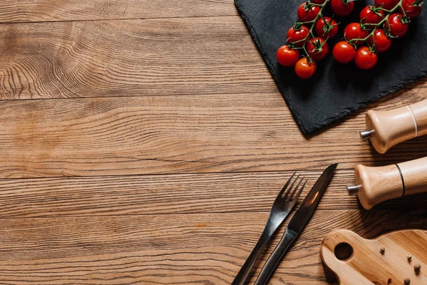 Top view of fresh ripe tomatoes on slate board, fork with knife, seasonings in containers and peppercorns on wooden table — Stock Photo