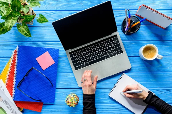 Geschäftsmann schreibt in Notizblock per Laptop auf blauem Holztisch mit Schreibwaren — Stockfoto