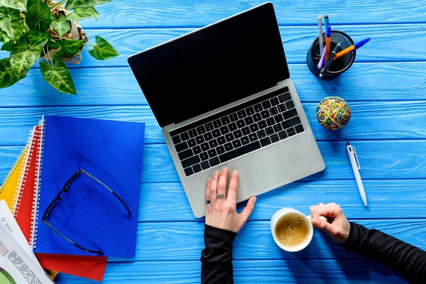 Hand mit Kaffeetasse per Laptop auf blauem Holztisch mit Schreibwaren — Stockfoto