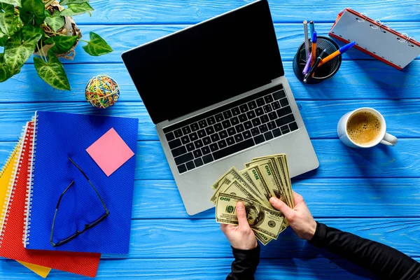Деловой человек считает доллары на ноутбуке на синем деревянном столе с канцелярскими принадлежностями — стоковое фото