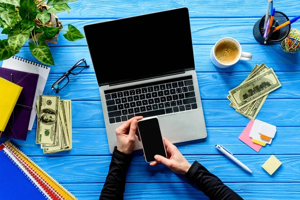 Крупным планом человека, использующего смартфон на ноутбуке на синем деревянном столе с канцелярскими принадлежностями и деньгами — стоковое фото