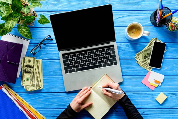 Person writing in notebook by laptop on blue wooden table with stationery and money — Stock Photo