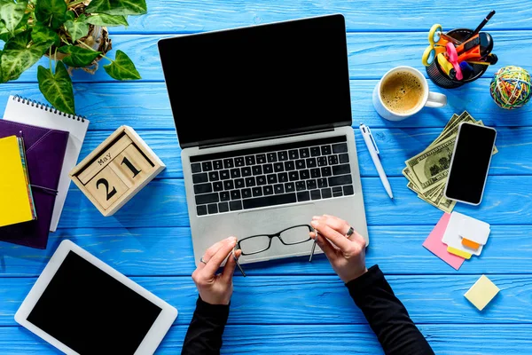 Manos que sostienen las gafas por el ordenador portátil en la mesa azul de madera con el papel y el dinero - foto de stock