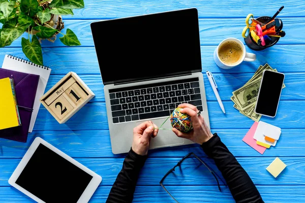 Деловой человек привязывает резиновые ленты к мячу на ноутбуке на синем деревянном столе с канцелярскими принадлежностями и деньгами — стоковое фото