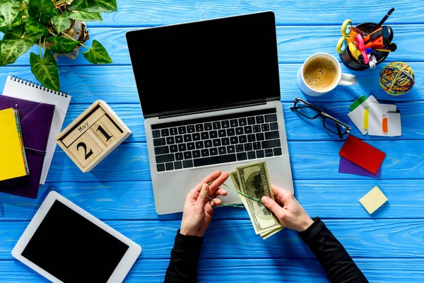 Business person tying dollars with rubber band by laptop on blue wooden table with stationery — Stock Photo