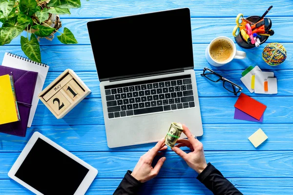 Geschäftsmann hält Dollars per Laptop auf blauem Holztisch mit Schreibwaren — Stockfoto