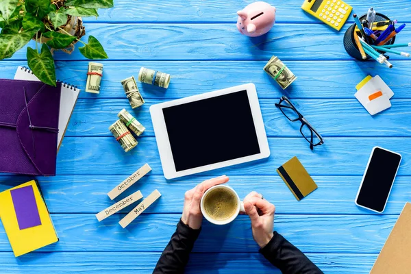 Mãos segurando xícara de café na mesa de madeira azul por dinheiro e tablet digital — Fotografia de Stock
