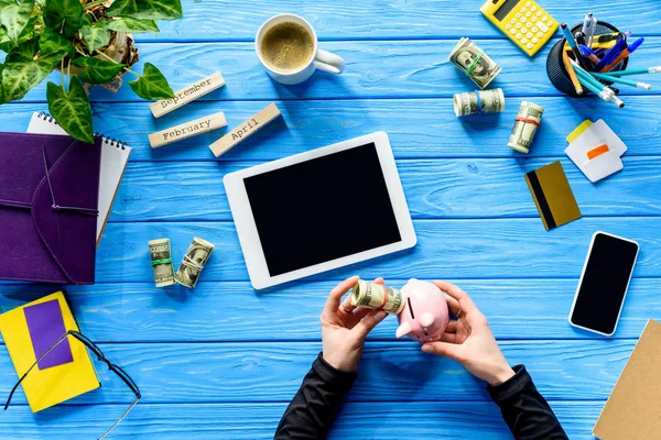 Mani che tengono salvadanaio e dollari su tavolo di legno blu con tablet — Foto stock