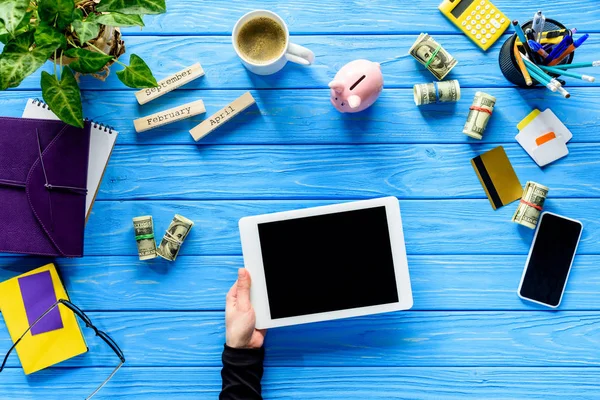 Primer plano de la mano sosteniendo la tableta digital en la mesa de madera azul con dinero y papelería - foto de stock