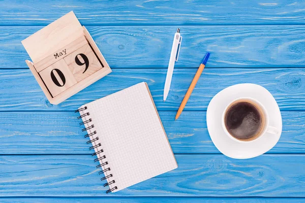 Draufsicht auf Holzkalender mit Datum 9. Mai, Kaffeetasse, leeres Lehrbuch und Kugelschreiber, Siegertagskonzept — Stockfoto