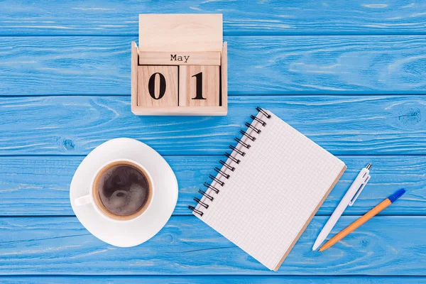 Vista superior del calendario de madera con fecha del 1 de mayo, taza de café, libro de texto en blanco y bolígrafos, concepto de día de los trabajadores internacionales - foto de stock