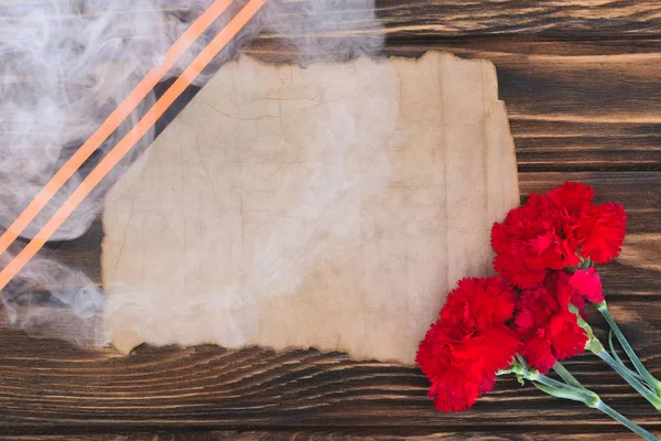 Smoke over st. george ribbon, old empty paper and carnations on wooden surface — Stock Photo