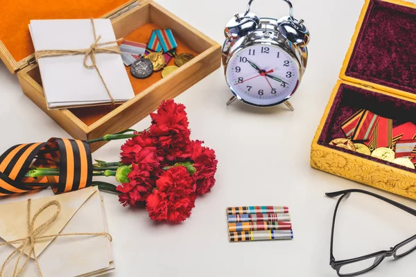 Primer plano de despertador, anteojos, flores envueltas por la cinta de San Jorge, letras, medallas en gris, concepto del día de la victoria - foto de stock