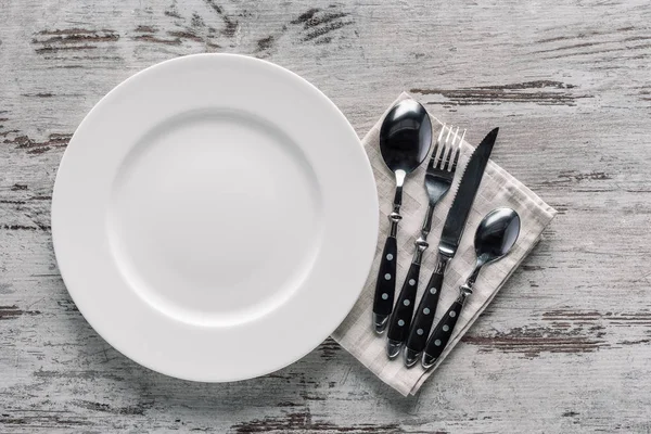 Белая тарелка и столовые приборы на салфетке на деревянном столе — стоковое фото