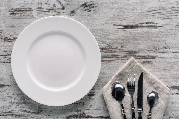 Белая тарелка и столовые приборы на салфетке на деревянном фоне — стоковое фото