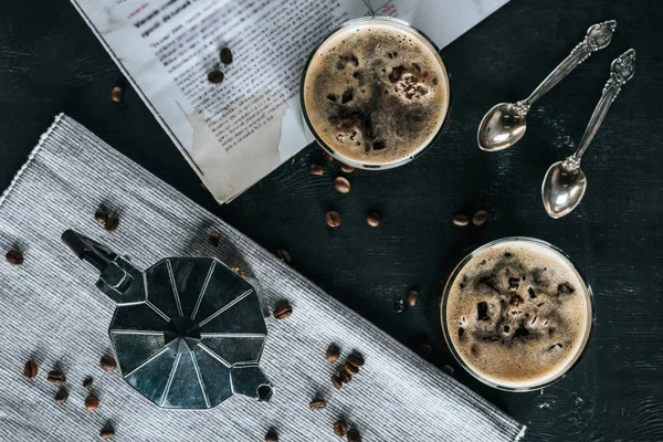 Flat lay com cafeteira, colheres, jornal e copos de café frio em mesa preta — Fotografia de Stock