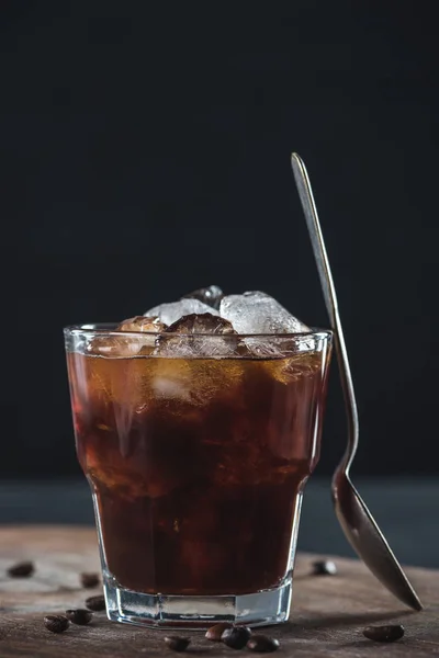 Close up view of glass of cold brewed coffee with spoon on wooden cutting board on dark backdrop — Stock Photo