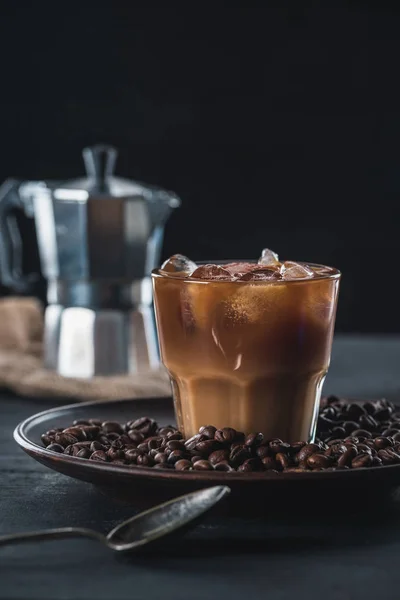 Enfoque selectivo de vaso de café helado frío en el plato con frijoles tostados y cafetera en la mesa sobre fondo oscuro — Stock Photo